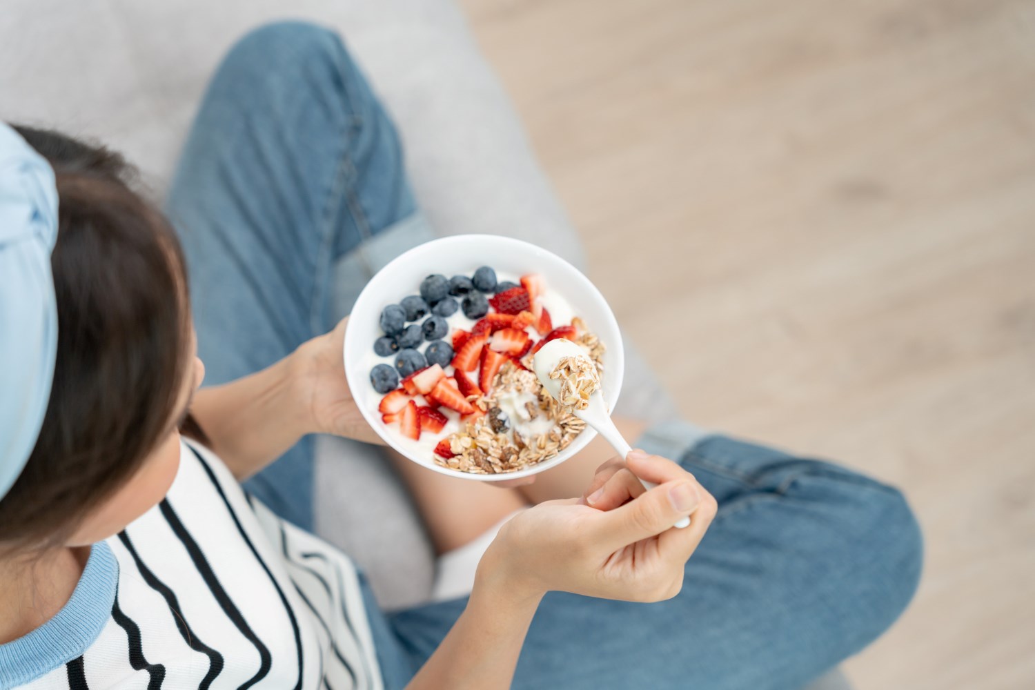 Woman in striped t-shirt sat on a wooden floor with a spoon and granola and fruit bowl in her hands